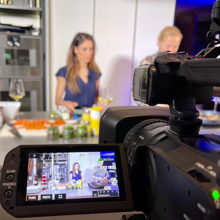 Zwei Personen in einer Küche bei der Zubereitung von Speisen, Kameraaufnahme im Vordergrund.